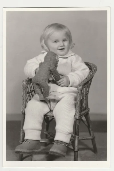 Foto vintage muestra una pequeña viga se sienta en la silla de mimbre y sostiene el juguete de peluche (peluche). Fotografía en blanco y negro retro . — Foto de Stock