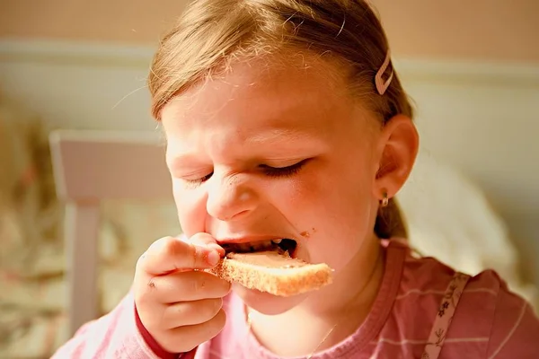 Una linda niña pequeña. Una niña pequeña come un brindis con crema de chocolate. Una niña pequeña se sienta en la cocina . —  Fotos de Stock