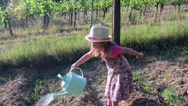 Niña lleva vestido floral y sombrero blanco. Niña riega el jardín — Vídeo de stock