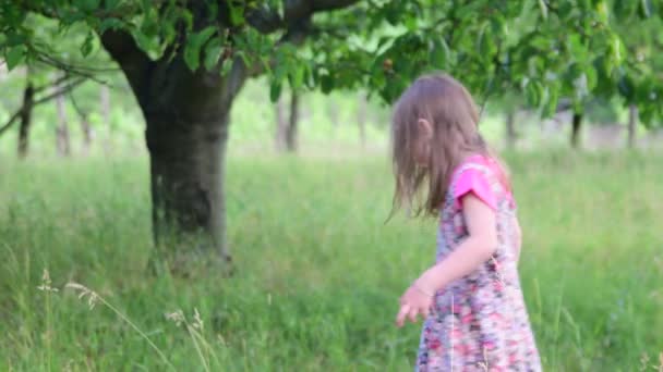 Мила дівчина танцює в природному саду. Маленька дівчинка танцює і стрибає на маленькому батуті. Маленька дівчинка носить квіткову сукню та білий капелюх — стокове відео