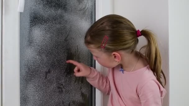 La fille dessine un doigt sur une vitre. Petite fille mignonne essuie fenêtre gelée en hiver . — Video