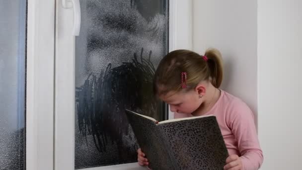 Niña pequeña se sienta en el alféizar de la ventana detrás de la ventana congelada. Linda niña se sienta en la repisa de la ventana y lee el libro . — Vídeos de Stock