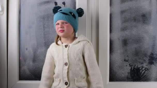 Niña pequeña se sienta en el alféizar de la ventana detrás de la ventana congelada . — Vídeos de Stock