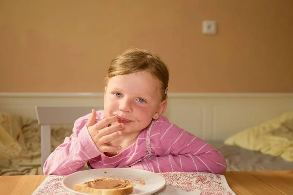 La chica feliz tiene un aperitivo en la cocina. Una linda niña pequeña sonríe. Una niña pequeña con manchas de crema de chocolate en la cara. Una niña pequeña se sienta en la cocina . —  Fotos de Stock