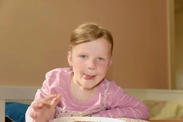 La chica feliz tiene un aperitivo en la cocina. Una linda niña pequeña sonríe. Una niña pequeña con manchas de crema de chocolate en la cara. Concepto de infancia y diversión . —  Fotos de Stock