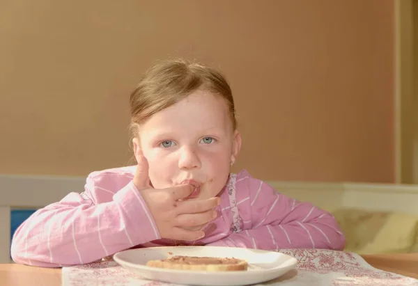 Šťastná dívka má svačinu v kuchyni. Roztomilá malá dívka úsměvy. Malá dívka s čokoládový krém skvrny na obličeji. Koncept dětství a zábavy. — Stock fotografie