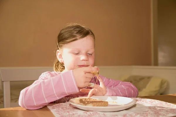 Niña juguetona come crema de chocolate untada en el pan. Snack de comida dulce de chocolate. La chica feliz tiene un aperitivo en la cocina. Una linda niña pequeña sonríe. Una niña pequeña con manchas de crema de chocolate en la cara —  Fotos de Stock