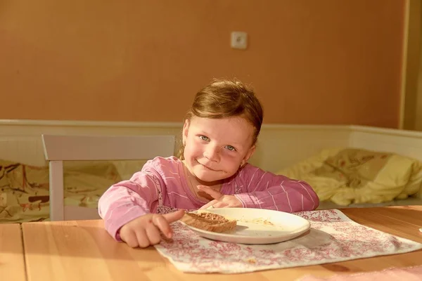 Девочка-игривая ест шоколадное мороженое, намазанное на хлеб. Шоколадная закуска. Счастливая девушка перекусила на кухне. Милая маленькая девочка улыбается. Маленькая девочка с шоколадными пятнами на лице — стоковое фото