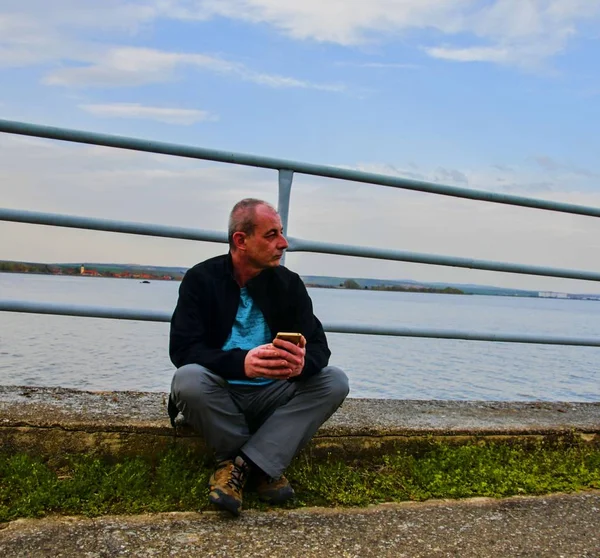 Homem de meia idade sentado na margem do lago. Solidão homem maduro usando telefone celular no banco. Conceito de homem activo — Fotografia de Stock