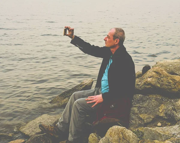 Belterület éves ember ül a tó partján. Magány érett férfi figyelembe selfie kép a parton. Magány fogalmának — Stock Fotó