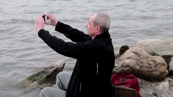 Hombre de mediana edad sentado en la orilla del lago. Soledad hombre maduro tomando foto selfie en el banco. Concepto de soledad — Vídeos de Stock