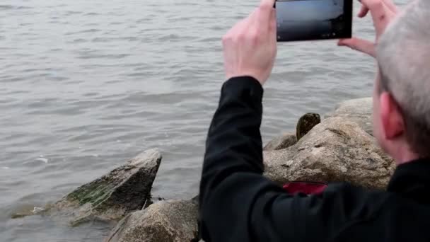 Zijaanzicht leeftijd man zit op de oever van het meer. Eenzaamheid volwassen man schieten video op de bank. Concept van eenzaamheid — Stockvideo