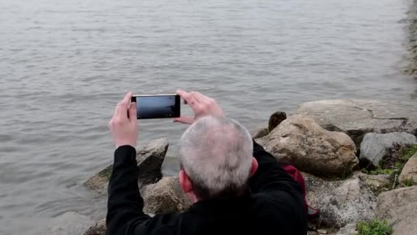 Midle wieku człowiek siedzi na brzegu jeziora. Samotność dojrzałe man nagrywania wideo przez telefon komórkowy na bank. Pojęcie samotności — Wideo stockowe