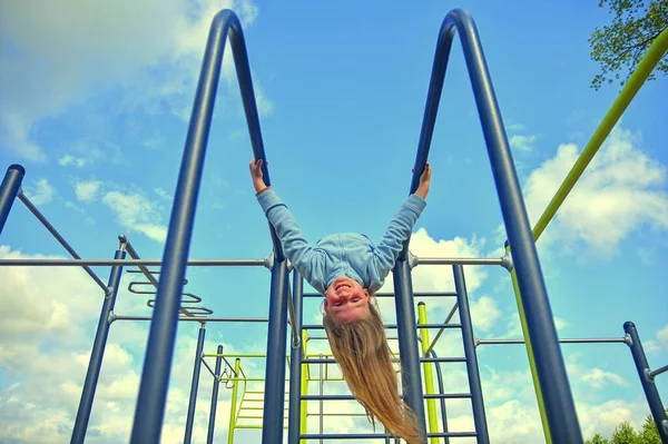 Niski kąt widzenia szczęśliwej dziewczyny wiszącej na ramie wspinaczkowej na placu zabaw patrzącej na kamerę uśmiechniętą i cieszącą się latem. — Zdjęcie stockowe