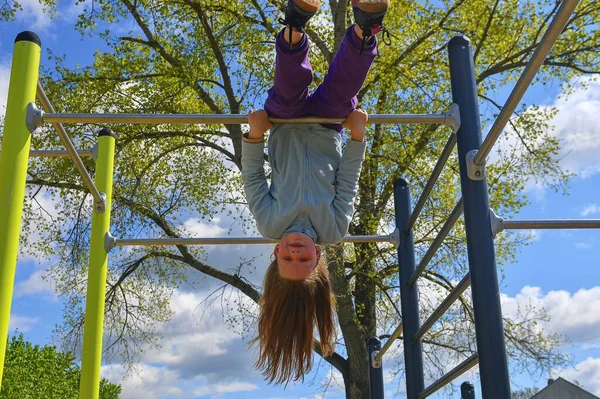 Urocza dziewczynka śmiejąca się szczęśliwie, wisząca do góry nogami na równoległych barach na placu zabaw. — Zdjęcie stockowe