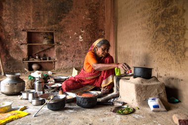 CHIKHALDARA, MAHARASHTRA - AUGUST 28, 2018: An Unidentified old woman making and cooking / baking fresh food in a rural village in a vintage kitchen using firewood in earthen chulhas. clipart