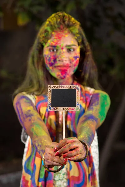 粉末色で描かれた顔を持つホリ祭りの際に小さなボードを保持する若い女の子 色のスプラッシュと — ストック写真