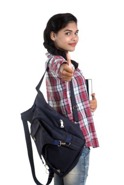 Sırt çantalı genç Hintli kadın ayakta duruyor ve elinde defterlerle beyaz bir arka planda poz veriyor..