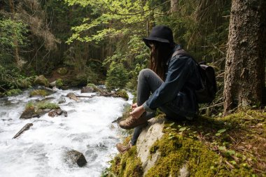 Genç, yalnız, tatlı turist kız dağların kıyısında taşın üzerinde oturuyor vahşi arazide ve susuzluğunu gidermek için bardaktan su içiyor. Gezgin doğada dinleniyor. Sıvı dengesi. Sıcaklık