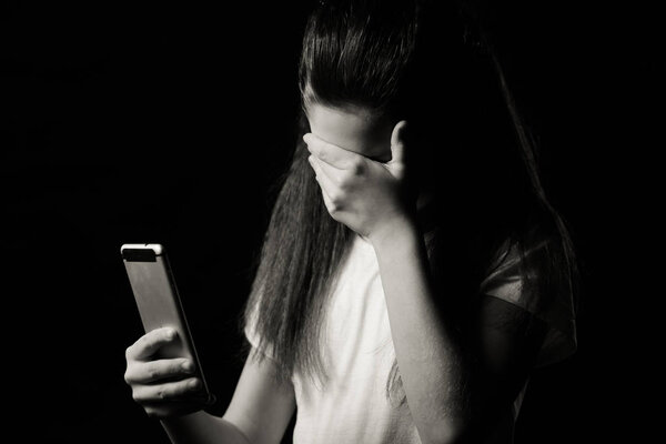 Single sad teen holding a mobile phone lamenting sitting on the bed in her bedroom with a dark light in the background