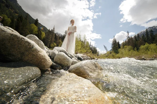Novia feliz en un vestido blanco, una corona de cachorros vivos y hojas y un ramo de flores en la orilla de un río de montaña —  Fotos de Stock