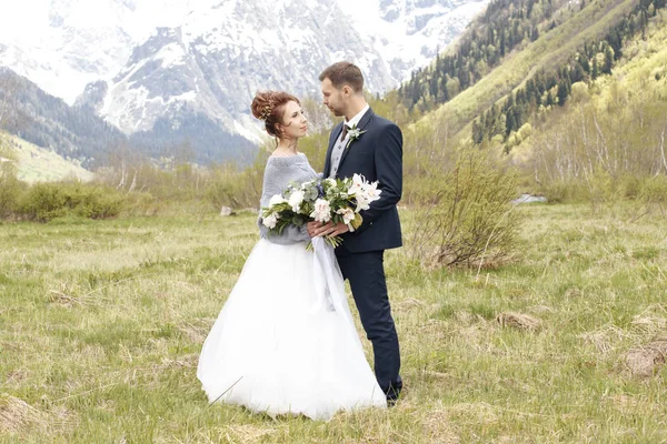 Una pareja en un vestido de novia con un ramo de flores y verduras está en las manos en el fondo de las montañas al atardecer, la novia y el novio —  Fotos de Stock