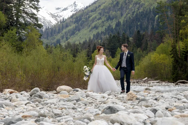 Paseo nupcial. La novia y el novio caminan a lo largo de la orilla del río, cogiéndose de las manos. Retrato de los novios. En el fondo del lago . —  Fotos de Stock