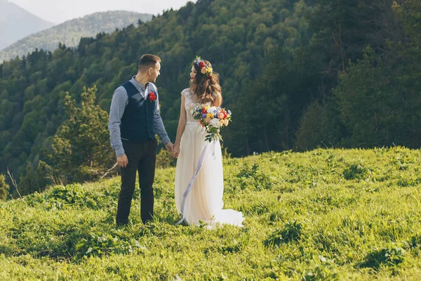 Pareja en traje de novia con un ramo de flores y vegetación está en las manos contra el telón de fondo del campo al atardecer, la novia y el novio —  Fotos de Stock