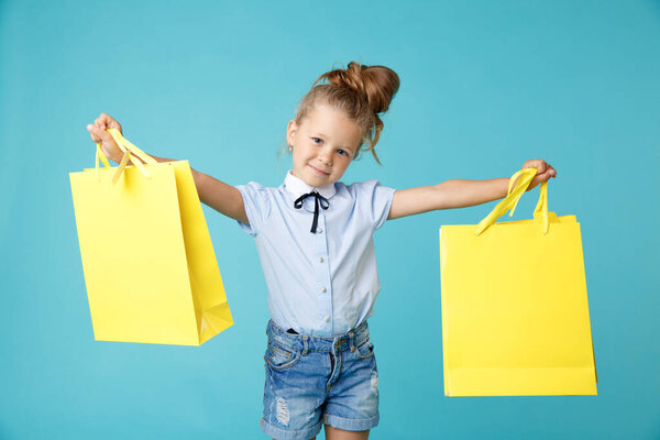 Little cute and funny girl holding big bright colored yellow paper bags in the blue studio.