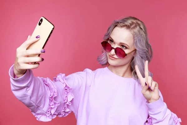 Vtipné dívka v brýlích dělat vlastní fotografii na svém telefonu v růžovém pokoji. — Stock fotografie