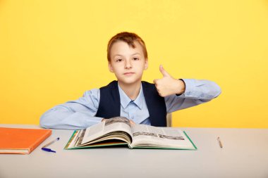 Çocuk masada oturuyor ve sarı arka planda baş parmak işareti gösteriyor..