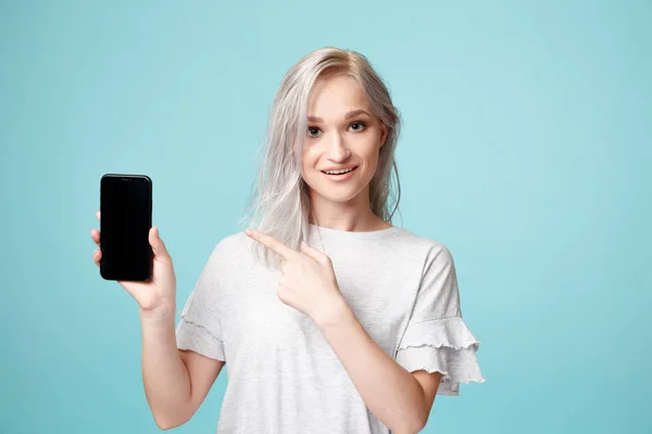 Veselá žena v T-Shurt ukazující na telefon v modrém studiu. — Stock fotografie