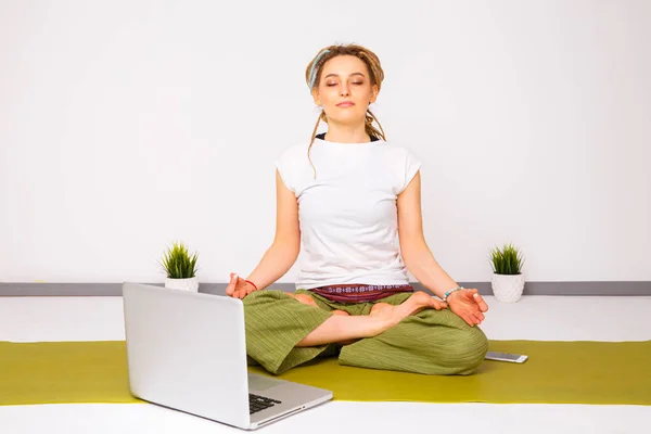 Woman yoga doing lotus pose on the green mat on the floor near the laptop.