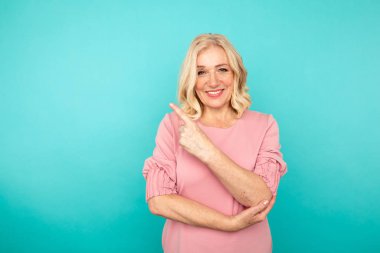 Yüzünde gülücükler açan orta boylu sarışın bir kadın mavi duvarda duran bir şeyi işaret ediyor..