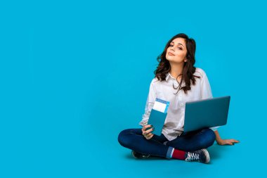 Young woman sitting on the floor with laptop and passport buying tichets online.