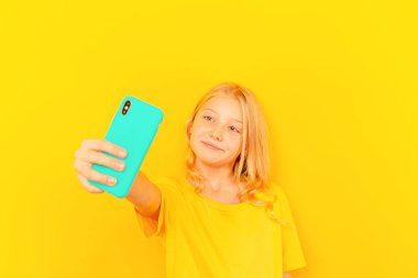 Gülümseyen küçük kız açık sarı arka planda yeni popüler cep telefonunun mavi ekranını gösteriyor..