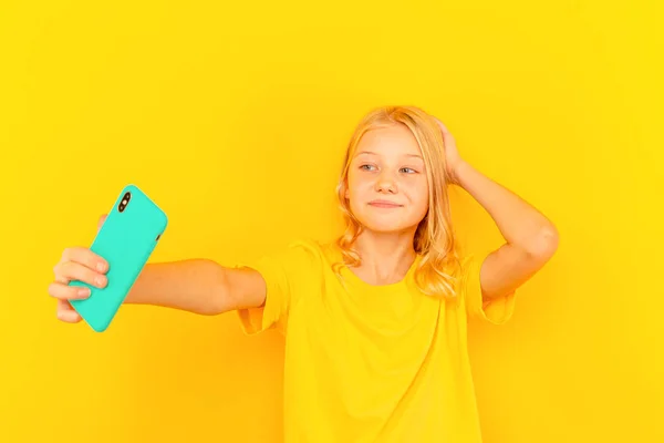 Usmívající se holčička dítě ukazující modré obrazovce nového populárního mobilního telefonu na světle žlutém pozadí. — Stock fotografie
