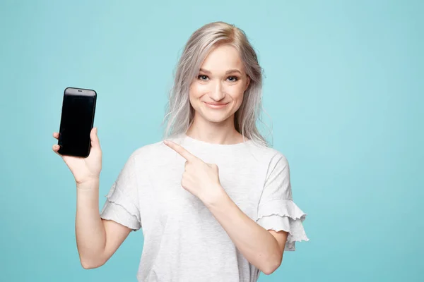 Veselá žena v T-Shurt ukazující na telefon v modrém studiu. — Stock fotografie