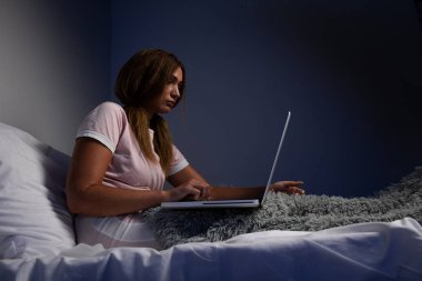 Depresyonda olan ciddi bir kadın yatakta oturuyor ve dizüstü bilgisayarla çalışıyor..