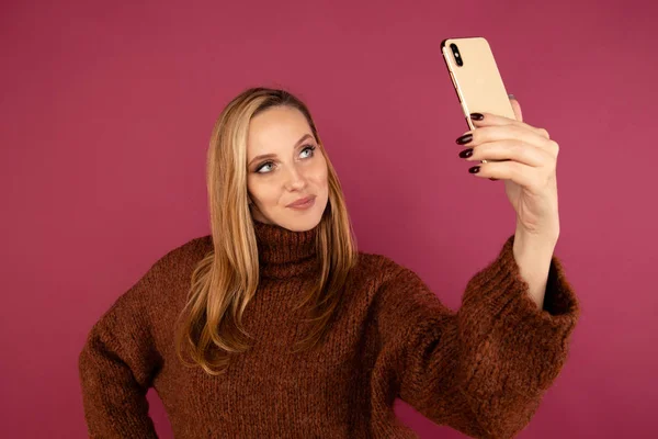 Mladá dáma ve svetru dělat self photo na telefonu v růžovém studiu. — Stock fotografie