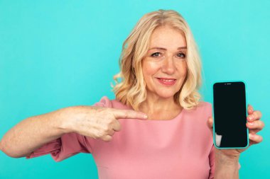 Happy smiling mature woman pointing phone in the blue studio.