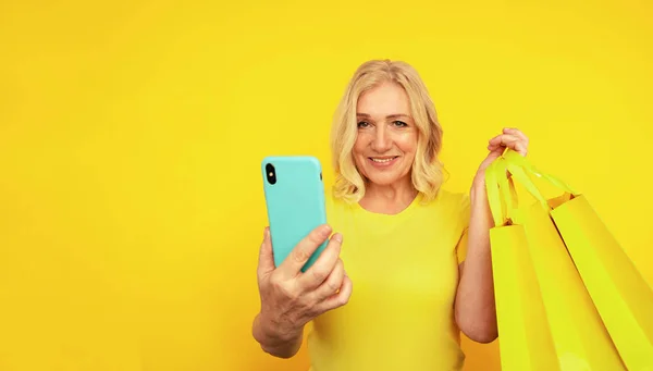 Gelukkig volwassen vrouw op de gele achtergrond poseren met tassen en heldere telefoon. — Stockfoto
