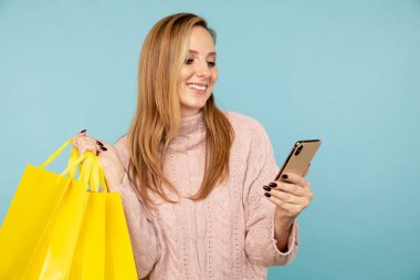 Gülümseyen ve telefon kullanan genç bir kadın sarı alışveriş torbaları taşıyor..