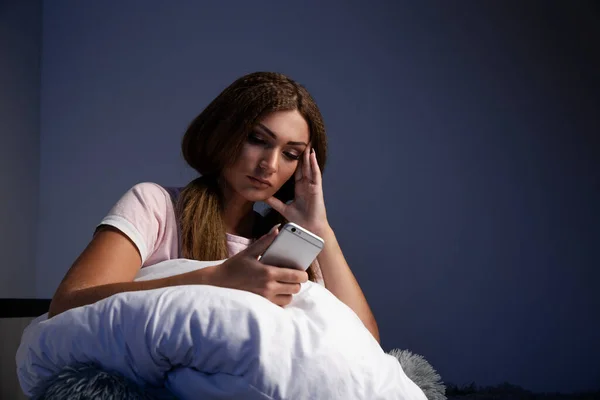 Mujer cansada en la cama usando teléfono sentado con almohada . —  Fotos de Stock