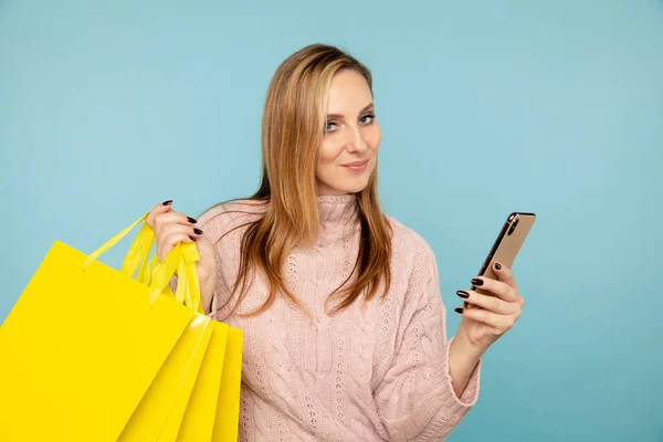 Hezká mladá žena se žlutými taškami a telefonem izolovaným na modrém pozadí. Koncept prodeje. — Stock fotografie