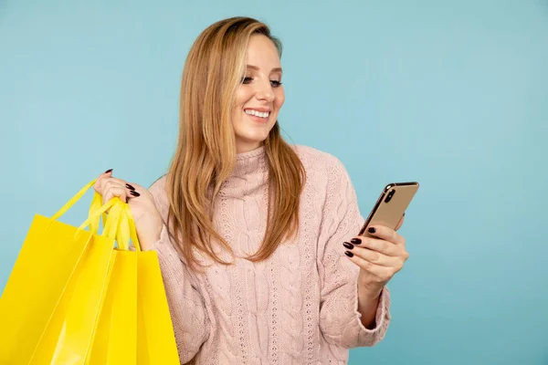 Jonge glimlachende vrouw glimlachen en met behulp van telefoon met gele boodschappentassen geïsoleerd. — Stockfoto