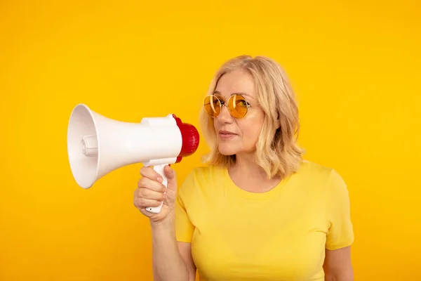 Leuke blonde mid vrouw spreken in de megafoon geïsoleerd over de gele achtergrond. — Stockfoto