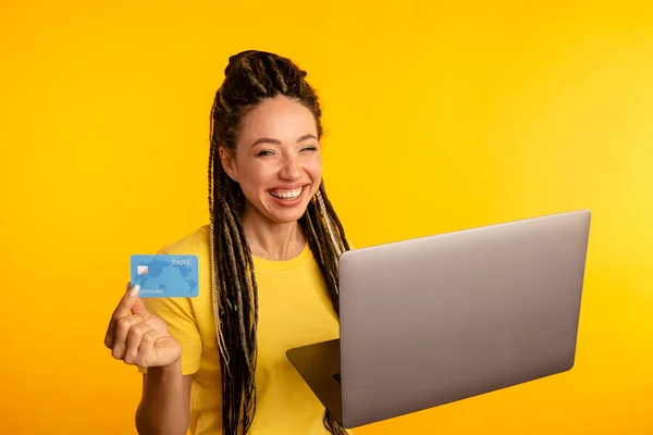 Portret szczęśliwej młodej damy z laptopem i kartą kredytową odizolowanej od żółtego studia. — Zdjęcie stockowe