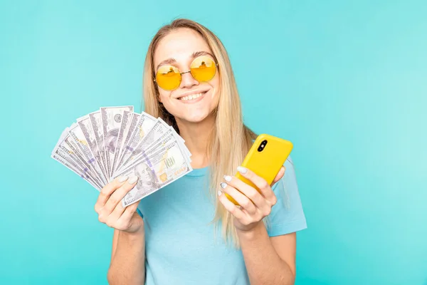 Jovem senhora isolado sobre fundo azul. Olhando câmera mostrando exibição de telefone celular segurando dinheiro . — Fotografia de Stock