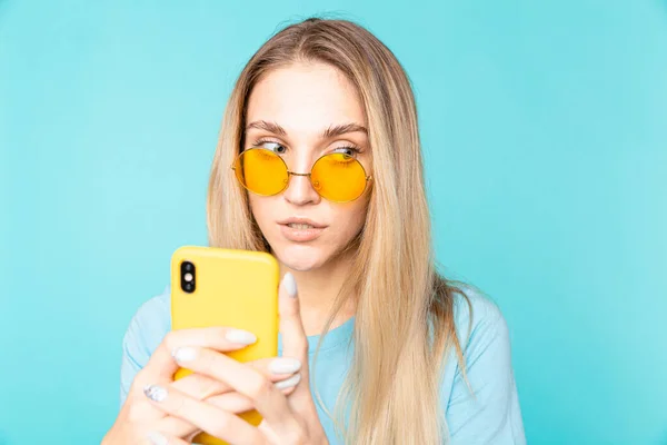 Imagine a unei tinere blonde care pozează izolată pe fundalul peretelui albastru folosind telefonul mobil — Fotografie, imagine de stoc